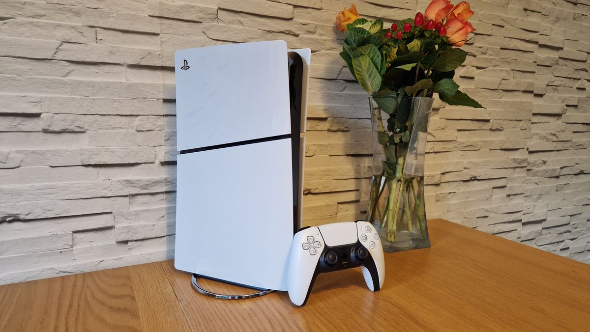 Console PS5 Slim e controller DualSense su un tavolo di legno di fronte a uno sfondo di mattoni bianchi e accanto a un vaso di fiori